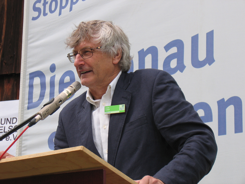 Ein grauhaariger Mann mit Brille und einem grünen Anstecker am dunklen Sakko spricht an einem Rednerpult ins Mikrophon. Hinter ihm ein beschriftetes Banner. (Foto: BN)