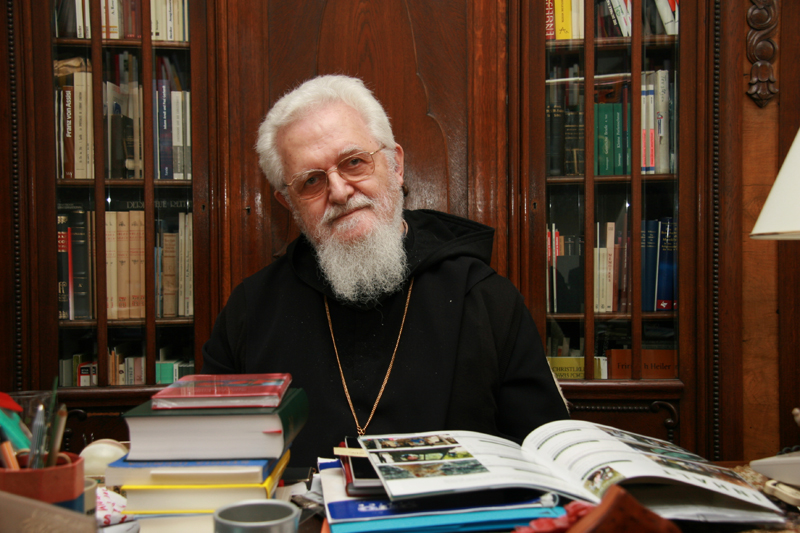Ein weißhaariger Mann mit Brille und Bart, einer Mönchskutte und einer Kette um den Hals sitzt hinter einem Tisch mit vielen Büchern, im Hintergrund Bücherschränke (Foto: Paulus)