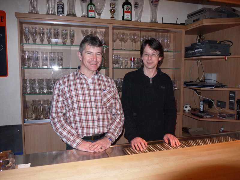 Zwei Männer, einer mit kariertem Hemd der andere mit einem dunklen Pullover, stehen hinter einem Schanktisch, hinter ihnen Regale mit Gläsern (Foto: BN) 