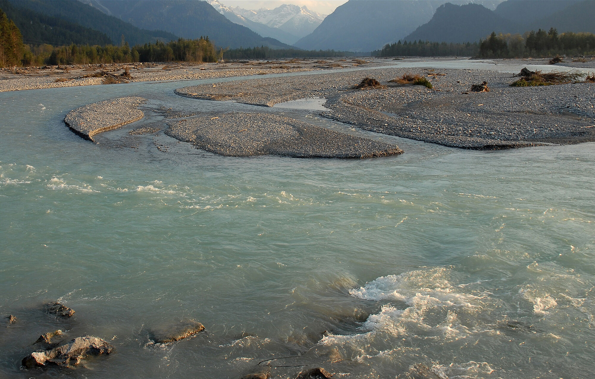 Ein Fluss mit gletscherblauem Wasser dehnt sich zwischen zahlreichen Sanbänken aus. An den Rändern wächst Wald, im Hintergrund Berge.