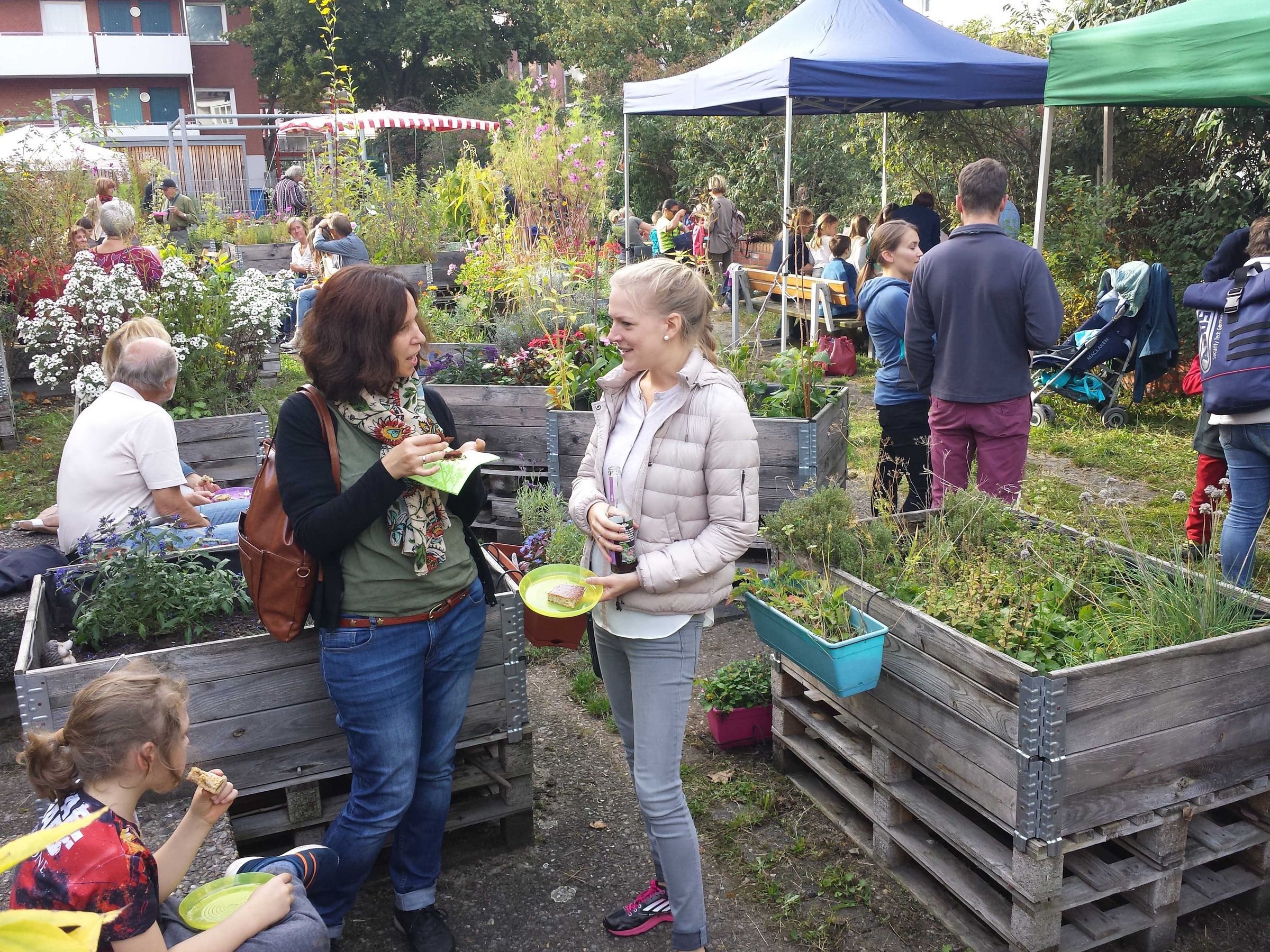 Urban Gardening: Menschen stehen mit Getränken und Kuchen in den Händen zwischen Hochbeeten und unterhalten sich. (Foto: Anne Tieseler)
