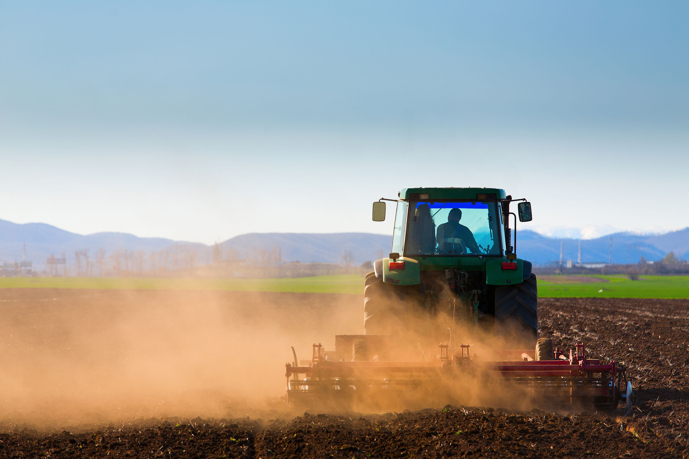 Ein Traktor fährt vor einer Bergkulisse über ein Stoppelfeld und wirbelt dabei viel rötlichen Staub auf. Äcker setzen Klimagase frei, deshalb hat Grünland eine große Bedeutung für den Klimaschutz. (Foto: danmir12/stock.adobe.com) 