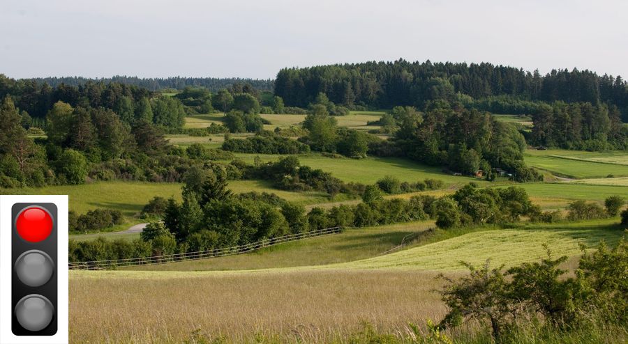 Eine hügelige Landschaft mit Wiesen und vielen Hecken und eine Ampel, die auf Rot steht. Bereits seit 2002 besteht im Bundesnaturschutzgesetz die Verpflichtung, auf 10 Prozent der Fläche jeden Bundeslandes einen Biotopverbund zu schaffen. (Foto: leonidp/stock.adobe.com, Ampel: kledesign/stock.adobe.com) 