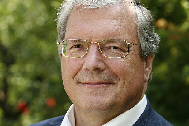 Das Portrait eines Mannes mit weißen Haaren und Brille in einem weißen Hemd und einer blauen Anzugjacke.