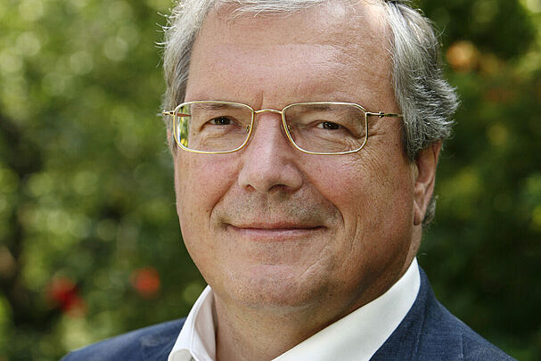 Das Portrait eines Mannes mit weißen Haaren und Brille in einem weißen Hemd und einer blauen Anzugjacke.