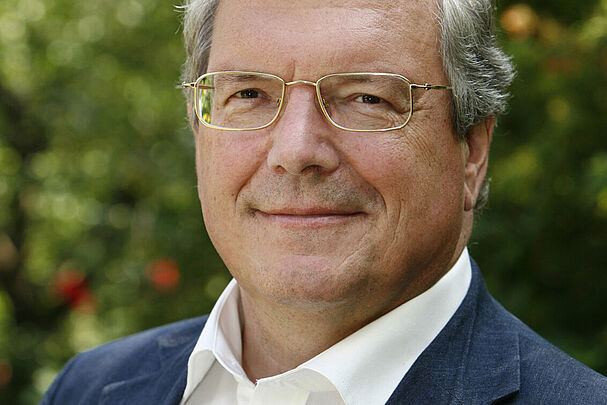 Das Portrait eines Mannes mit weißen Haaren und Brille in einem weißen Hemd und einer blauen Anzugjacke.