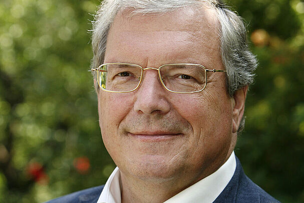 Das Portrait eines Mannes mit weißen Haaren und Brille in einem weißen Hemd und einer blauen Anzugjacke.