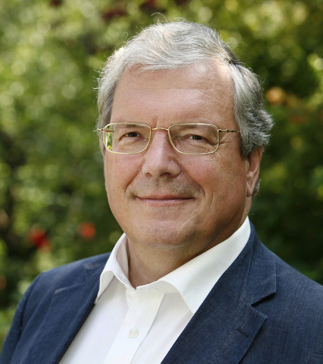 Das Portrait eines Mannes mit weißen Haaren und Brille in einem weißen Hemd und einer blauen Anzugjacke.