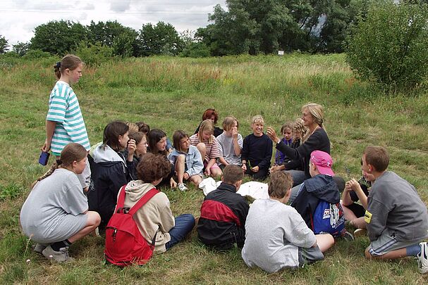 Kinder sitzen im Kreis, eine Frau hält eine Pflanze hoch und erklärt etwas. (Foto: Archiv SandAchse)