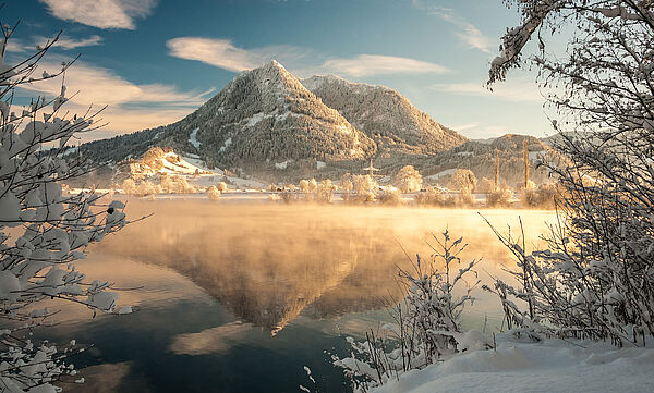 Der schneebedeckte Grünten im Winter erstrahlt in schrägem Sonnenlicht. Im Vordergrund liegt ein See, aus dem Nebel aufsteigt.