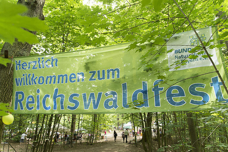 Im Wald hängt ein großes grünes Banner mit der Aufschrift "Herzlich willkommen zum Reichswaldfest" (Foto: Toni Mader)