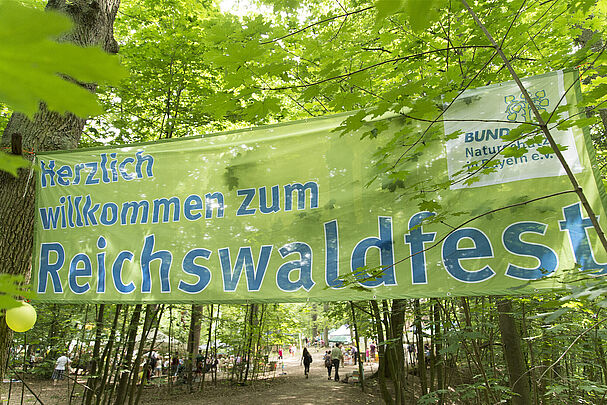 Im Wald hängt ein großes grünes Banner mit der Aufschrift "Herzlich willkommen zum Reichswaldfest" (Foto: Toni Mader)