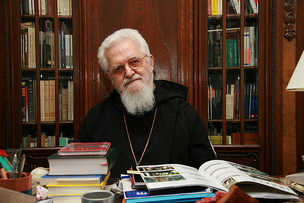 Ein weißhaariger Mann mit Brille und Bart, einer Mönchskutte und einer Kette um den Hals sitzt hinter einem Tisch mit vielen Büchern, im Hintergrund Bücherschränke (Foto: Paulus)