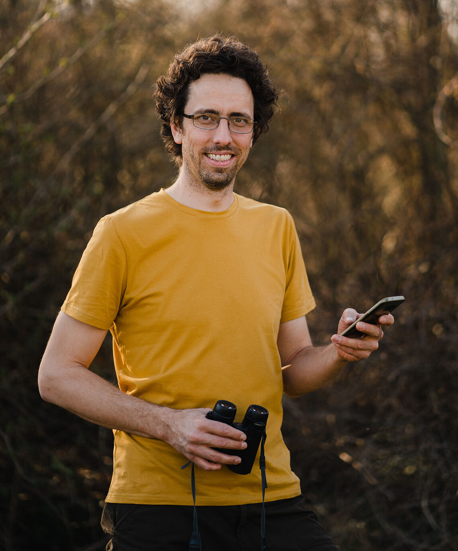 Ein Mann mit Brille und einem orangefarbenen T-Shirt hält ein Smartphone und eine Fernglas in der Hand. Philipp Herrmann, wie der „Vogelphilipp“ wirklich heißt, erkennt alle heimischen Singvögel an ihrer Stimme. (Foto: Alexey Testov) 