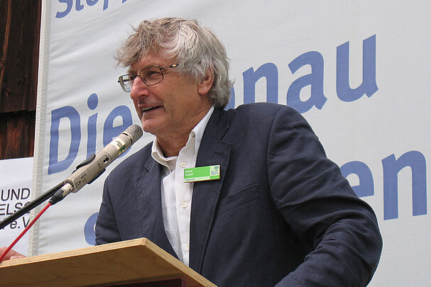 Ein grauhaariger Mann mit Brille und einem grünen Anstecker am dunklen Sakko spricht an einem Rednerpult ins Mikrophon. Hinter ihm ein beschriftetes Banner. (Foto: BN)