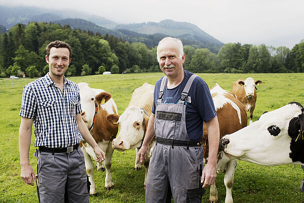 Junior- und Seniorlandwirt stehen in Arbeitskleidung auf der Weide bei ihren Kühen.
