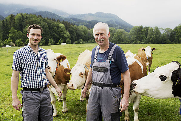 Junior- und Seniorlandwirt stehen in Arbeitskleidung auf der Weide bei ihren Kühen.