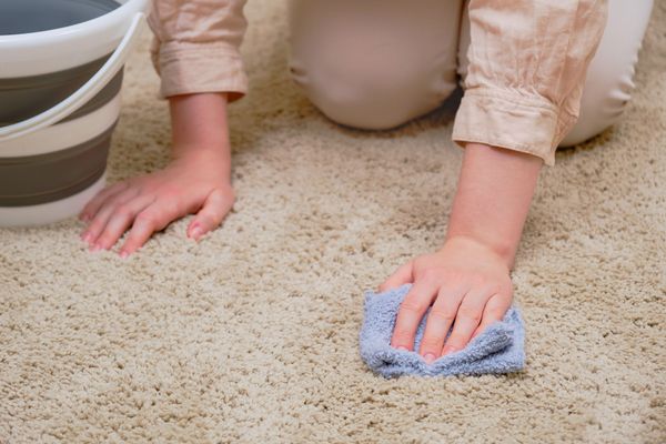 Eine Frau kniet mit einem Lappen in der Hand auf einem hellen Boden, neben ihr ein Eimer. Leichte Flecken entfernt man mit Wasser oder Waschmittel. 