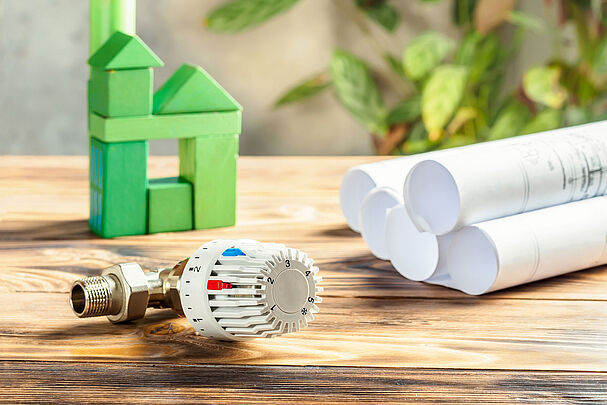 Ein Heizungsthermostat, ein Haus aus Bausteinen und einige Planungsrollen (Foto: IRINA/stock.adobe.com)