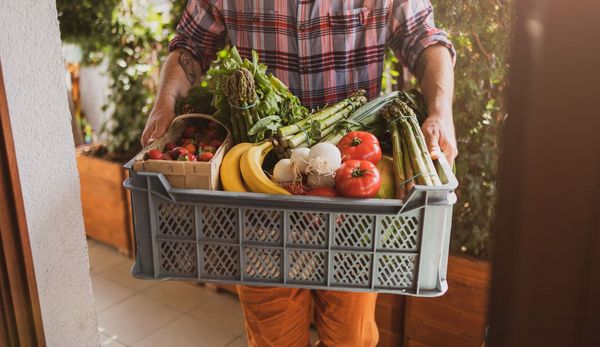 Ein Mann trägt eine offene Kiste mit verschiedenen Gemüsesorten in ein Haus.