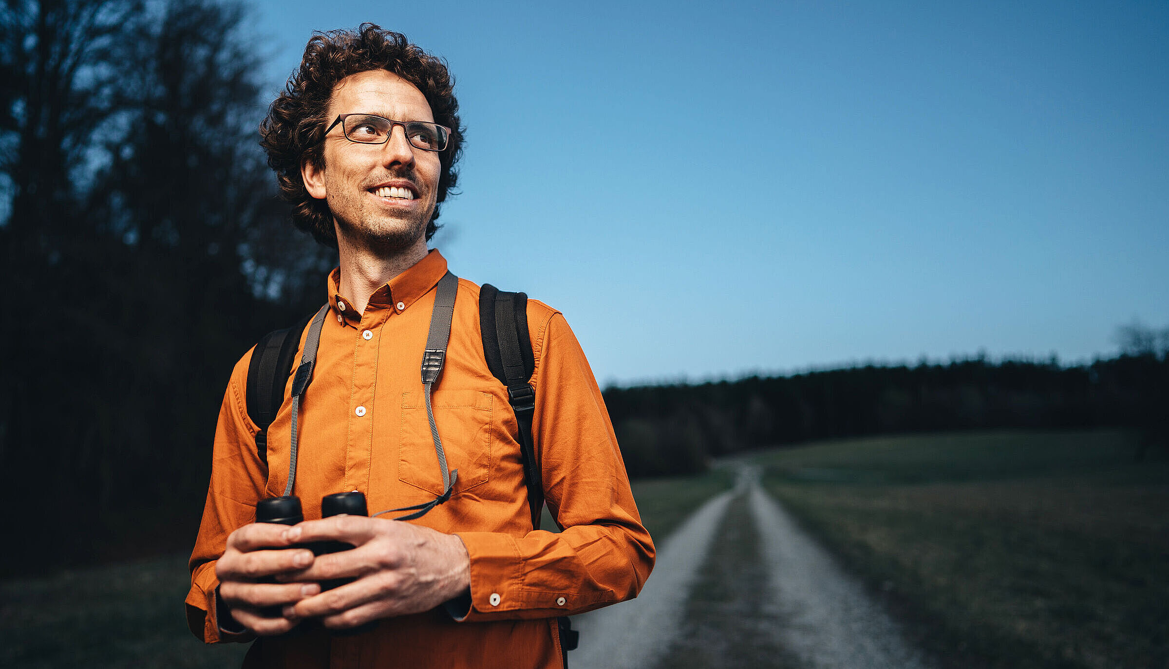 Ein Mann mit Brille und orangefarbenem Hemd steht auf einem Feldweg und hält ein Fernglas in der Hand. Der Vogelphilipp erkennt alle einheimischen Vögel an ihrem Gesang. (Foto: Alexey Testov) 