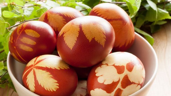 Ostereier natürlich färben: In einer weißen Schüssel liegen mehrere mit Zwiebelschalen gefärbte, rotbraune Eier mit hellen Pflanzenabdrücken auf der Schale.