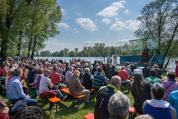 Menschen sitzen in Reihen auf Bierbänken und hören einem Redner auf einer Bühne zu. Im Hintergrund fließt die Donau. (Foto: Heinrich Inkoferer)