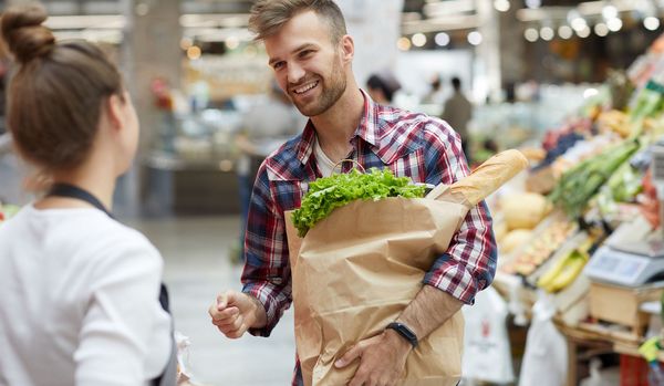 Lebensmittelverschwendung vermeiden: Ein junger Mann im Gespräch mit einer Verkäuferin vor einem Gemüseregal.