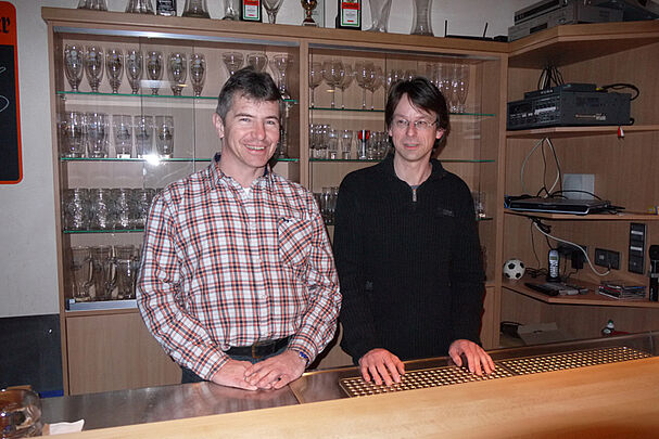 Zwei Männer, einer mit kariertem Hemd der andere mit einem dunklen Pullover, stehen hinter einem Schanktisch, hinter ihnen Regale mit Gläsern (Foto: BN)